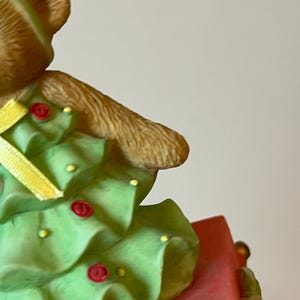 Puede incluir: Un oso de peluche marr&oacute;n que lleva un disfraz de &aacute;rbol de Navidad verde con una cinta amarilla y botones rojos. El oso est&aacute; sentado en una caja roja.