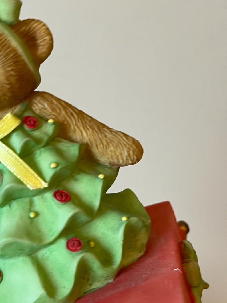 Puede incluir: Un oso de peluche marr&oacute;n que lleva un disfraz de &aacute;rbol de Navidad verde con una cinta amarilla y botones rojos. El oso est&aacute; sentado en una caja roja.