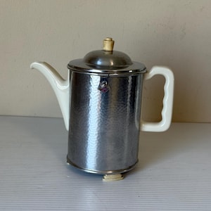 May include: A vintage silver-toned coffee pot with a hammered texture. It features a white ceramic handle, spout, and accents, and a small, light-colored knob on the lid. The pot is designed with a classic, retro aesthetic.