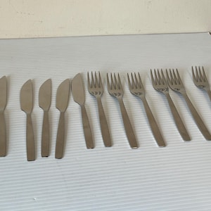 Puede incluir: Un juego de cubiertos de acero inoxidable, que incluye seis cuchillos y seis tenedores. Los cuchillos tienen una hoja curva y los tenedores tienen cuatro púas. Los cubiertos están colocados sobre una superficie blanca.