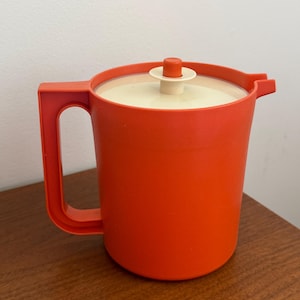 Può includere: Una caraffa in plastica arancione brillante con corpo cilindrico, una robusta maniglia laterale e un coperchio circolare color crema. Il coperchio include un piccolo pomello arancione centrale per la rimozione e un beccuccio sagomato.