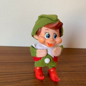 May include: A green and red elf doll with a white collar and a white pom-pom on its hat. The elf has brown hair, blue eyes, and a big smile.