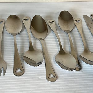 May include: A collection of stainless steel silverware, including forks, spoons, and knives. The spoons have a rounded bowl and a unique heart-shaped cutout at the end of the handle. The silverware is arranged on a white surface.