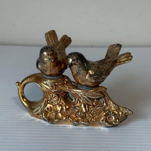 May include: A gold-coloured decorative piece featuring two birds perched on a floral branch. The birds are facing each other, and the base has a handle. The piece has an antique or vintage aesthetic.