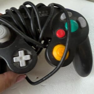 May include: A black Nintendo GameCube video game controller with a wrapped black cord. The controller features a grey directional pad, a yellow C-stick, a green A button, a red B button, and grey action buttons. The Nintendo GameCube logo is visible.