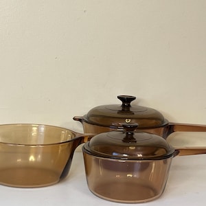 Puede incluir: Tres piezas de cristalería de cocina de color ámbar. El juego incluye dos cacerolas con tapa y mango largo y un cuenco abierto más grande. El cristal tiene un tono cálido y transparente.