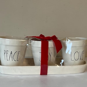 May include: A set of three off-white ceramic pots on a matching tray. Each pot has a word in black: PEACE, JOY, and LOVE. A red ribbon is tied around the middle pot. The set is wrapped in clear plastic.
