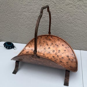 Peut inclure: Un porte-bûches en cuivre en forme de demi-cercle avec un motif floral texturé sur toute sa surface. L'objet comprend une haute poignée en métal arquée et repose sur quatre petits pieds décoratifs. Le métal présente une finition cuivrée.