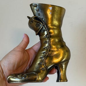 May include: A bronze-coloured decorative boot with a high heel and button details. The boot has a vintage style with a curved toe and a buckle detail on the side. The metallic finish gives it a classic, antique look.