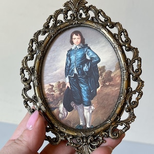 May include: An oval-shaped brass picture frame with an ornate design. The frame holds a vintage print of a young boy wearing a blue suit and holding a hat.