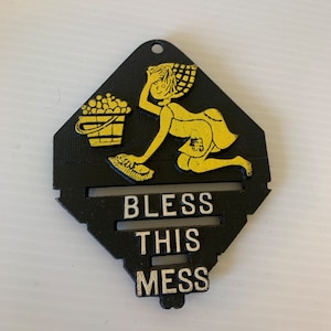 May include: Black and gold metal sign with a cartoon image of a woman holding a bucket and a broom. The sign reads "Bless This Mess".
