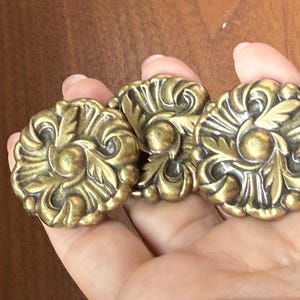 Könnte beinhalten: Drei runde, goldfarbene Schubladenknöpfe aus Metall mit einem erhabenen, komplexen Blumen- und Blattreliefmuster. Jeder Knopf hat eine kreisförmige Mitte, die von symmetrischen, geschwungenen Dekorelementen mit dunkler Antik-Optik umgeben ist.