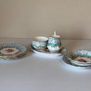 Puede incluir: Un juego de té vintage con un diseño floral turquesa y blanco. El juego incluye platos, platillos, una crema y una taza de té. Los platos y platillos tienen un borde dorado. El juego está dispuesto sobre una superficie blanca.