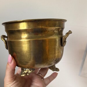 Peut inclure: Un petit pot rond en laiton métallique avec une finition polie, deux poignées latérales décoratives et trois pieds ornés. Le récipient présente une ouverture large, un corps lisse et incurvé, ainsi qu'une surface dorée aux nuances subtiles.