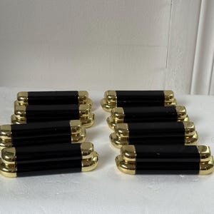 May include: A set of eight cabinet pulls featuring a rectangular black center section with rounded ends and polished gold-tone metal caps on both sides. The pulls are arranged in two columns of four, showcasing a smooth, reflective finish throughout.