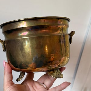May include: A polished brass pot with a rounded body, featuring two small decorative side handles and three ornate feet. The metal surface displays a warm, reflective golden-brown tone with subtle variations in colour and a smooth, metallic finish.