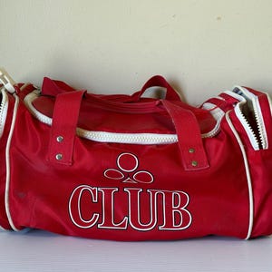 Puede incluir: Bolsa de deporte roja con ribete blanco y la palabra "CLUB" en letras blancas. La bolsa tiene una abertura con cremallera en cada extremo y una cremallera superior. Dos asas cortas están unidas a la parte superior de la bolsa.