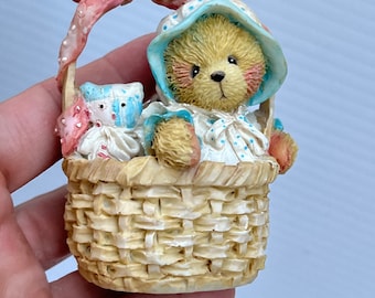 May include: A ceramic figurine of a teddy bear wearing a blue and white polka dot dress and a blue bonnet. The bear is sitting in a wicker basket with a pink ribbon handle. There is a smaller teddy bear in the basket with a pink and white polka dot dress.