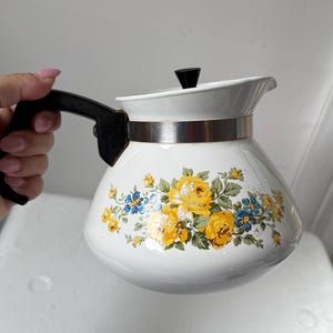 May include: A white enamel teapot featuring a floral pattern of yellow roses and small blue flowers. The teapot has a black plastic handle, a matching black knob on the lid, and a silver-toned metal band encircling the upper portion of the body.