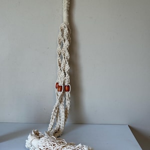 Peut inclure: Suspension de plante en macramé crème avec un anneau métallique pour accrocher. Le design comprend des nœuds tissés, des perles en bois et un bas à franges. Cet article fait main est conçu pour contenir une plante en pot.