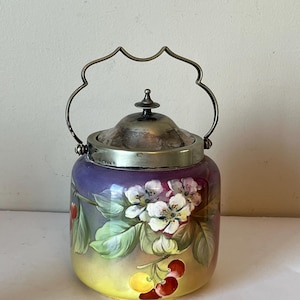 May include: A vintage ceramic cookie jar with a silver lid and handle. The jar is decorated with a floral design featuring white blossoms and red cherries on a purple and yellow background.