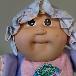 May include: A vintage Cabbage Patch Kid doll with a soft, vinyl face and brown eyes. The doll wears a lavender and pink outfit, including a bonnet with a floral pattern and a bib with the "Cabbage Patch Babyland" logo.