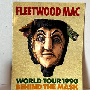 May include: A vintage concert poster for Fleetwood Mac's 1990 World Tour, "Behind the Mask". The poster features a colorful, wooden mask with a green snake and a bird on top.