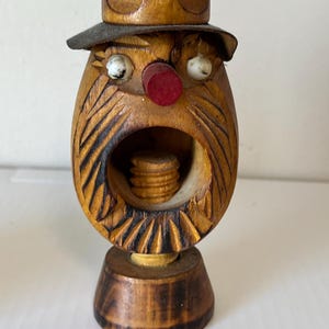 May include: A wooden nutcracker in the shape of a man's face with a hat. The nutcracker has a red nose, white eyes, and a carved beard. The nutcracker is made of light brown wood and has a dark brown hat.