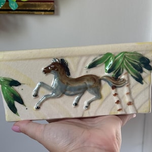 May include: A rectangular ceramic planter with a cream-coloured crackle glaze finish. The front features a raised, three-dimensional relief of a brown and grey horse galloping past green palm leaves and a stylised tree trunk against a textured background.