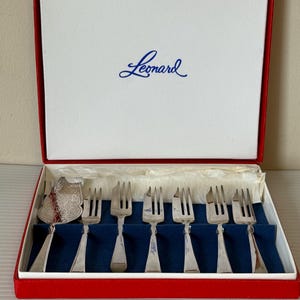 May include: A set of silver forks and a serving utensil in a red and white box with the name "Leonard" in blue script. The forks are arranged on a blue velvet-like surface.