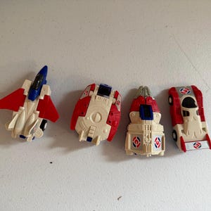May include: Four small plastic toy vehicles in a red, white, and blue color scheme. The set includes a fighter jet, a rounded spacecraft, a pointed shuttle-like craft, and a race car, each featuring circular emblems with red and blue markings.