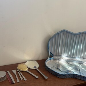 May include: A vanity set featuring a silver-toned hand mirror, a hairbrush with light-colored bristles, a comb, and three small metal tools. A round silver container is included, all presented with a blue patterned case lined with light blue satin.