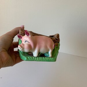 以下が含まれることがあります： 茶色と緑色のアクセントが付いたピンクのセラミック製のブタのプランター。ブタは緑色の土台の上に立っており、茶色の丸太で囲まれています。