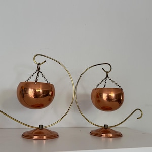 May include: Two vintage copper hanging planters. Each planter is a shiny copper bowl suspended by a chain from a curved brass-toned metal stand. The stands have a circular base and a decorative, curved design.
