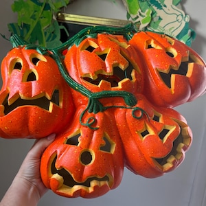 May include: A decorative wall hanging featuring four orange jack-o'-lantern pumpkins connected by green vines. Each pumpkin has carved facial features with cut-out eyes, noses, and jagged mouths. The surface has a speckled texture throughout.