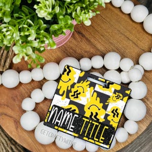 May include: A black and yellow badge holder with a checkerboard pattern and melting smiley faces. The badge holder has the words "NAME TITLE" printed on it. The background includes a wooden tray, white beads, and greenery.