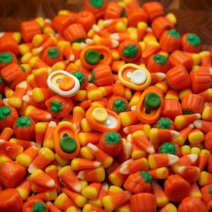 May include: A large pile of orange, yellow, and white candy corn and orange pumpkin-shaped candies. There are five small, colorful, plastic fidget toys on top of the candy.
