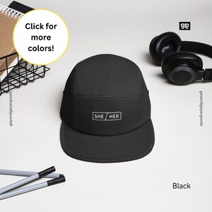 May include: Black 5-panel baseball cap with a white embroidered patch that says "SHE/HER".