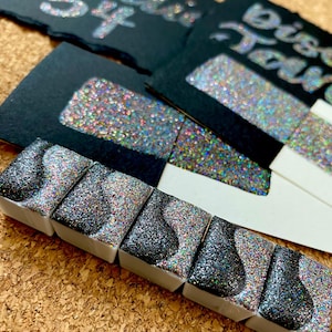 May include: Six small pans of black and silver glitter paint with a rainbow shimmer. The pans are arranged in a row on a cork surface. The paint is labeled "Black Galaxy".