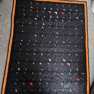 May include: A black quilt with a repeating pattern of colorful triangles and white dots. The quilt has an orange border and is likely handmade. The triangles are in various colors, including red, yellow, and purple.