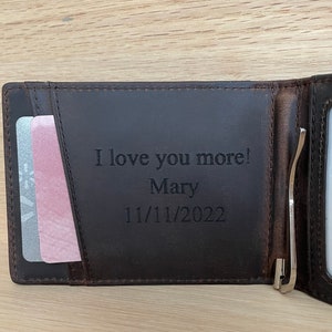 May include: Brown leather wallet with a money clip and a card slot. The wallet is engraved with the text "I love you more! Mary 11/11/2022".