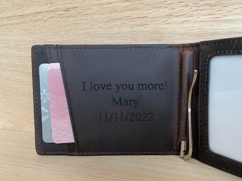 May include: Brown leather wallet with a money clip and a card slot. The wallet is engraved with the text "I love you more! Mary 11/11/2022".