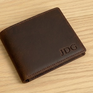 May include: A dark brown leather wallet with the initials "JDG" embossed on the front. The wallet is rectangular with rounded corners and visible stitching around the edges. The wallet is on a light brown wooden surface.