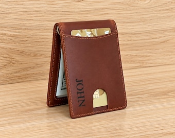 May include: A brown leather bifold wallet featuring a vertical money clip, a card slot with a thumb cutout, and the name JOHN embossed in black lettering on the front. The wallet holds a gold-colored credit card and a stack of paper currency.