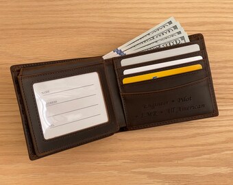 May include: A brown leather bifold wallet featuring a clear ID window with printed lines for name, address, and telephone, plus card slots. The wallet is engraved with the text Engineer, Pilot, EMR, and All American, and contains US currency.