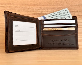 May include: Open brown leather wallet with several $100 bills visible. The wallet has slots for cards and a clear window for identification. The text "Engineer • Pilot • EMR • All American" is embossed on the inside.