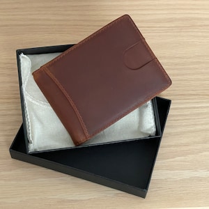 May include: A brown leather wallet in a black box with a white fabric lining. The wallet has a flap closure and a smooth leather finish.