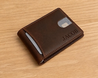 May include: A dark brown leather bifold wallet featuring visible stitching along the edges. The front exterior displays the name JACOB embossed in capital letters and includes a clear plastic window slot containing a card with a metallic chip.