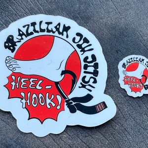 Könnte beinhalten: Zwei weiße Aufkleber mit einem roten Kreis und schwarzem Text. Der größere Aufkleber sagt "Brazilian Jiu Jitsu" und "Heel Hook!" mit einem Cartoon-Fuß und einem schwarzen Gürtel. Der kleinere Aufkleber ist das gleiche Design, aber kleiner.