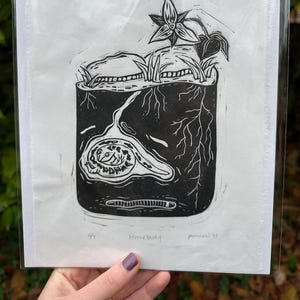 May include: Black and white linocut print of a plant in a pot, titled "Homebody". The artwork features a detailed illustration of a plant with roots, leaves, and a flower. The print is signed and numbered.